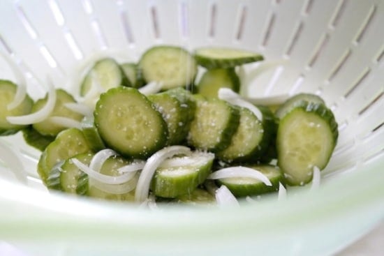 Sliced cucumbers and onions in a colander being salted