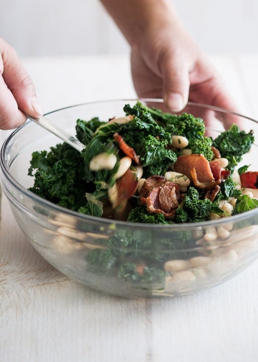 Kale, bacon and white bean salad being tossed in a glass bowl